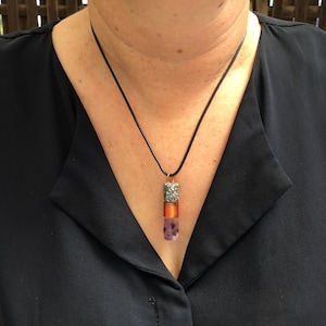 Op de afbeelding: Een ketting met hanger bestaande uit een zwart koord en een cilindervormige bedel. De bedel heeft een textuurrijk zilverkleurig bovendeel, een middendeel omwikkeld met koperkleurig draad en een gladde, lichtpaarse en witte stenen basis.