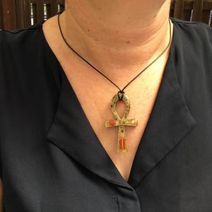 May include: An ankh pendant necklace featuring a translucent, speckled resin base with embedded orange and white stone accents and a small copper coil detail, suspended from a simple black cord that rests against a dark navy blue sleeveless top.