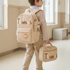 May include: A beige corduroy backpack and matching lunch bag set, both personalised with the name Thomas in brown and white embroidered lettering. The backpack features a small lion patch on the front pocket and a side pouch for a pencil case.