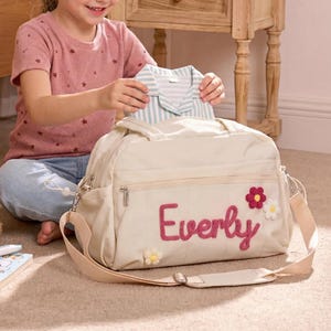 K&ouml;nnte beinhalten: Eine beige Wickeltasche mit dem Namen "Everly" in rosa Schrift und floralen Akzenten. Die Tasche hat einen Schultergurt und ist offen, mit einem gestreiften Hemd darin. Eine junge Person h&auml;lt das Hemd. Ein Teddyb&auml;r und ein Buch liegen auf dem Boden.
