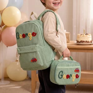 May include: A light green corduroy backpack and matching lunch bag set. The backpack and lunch bag have the name "George" embroidered in colourful letters. A small red car applique is on each bag. The bags are designed for children.