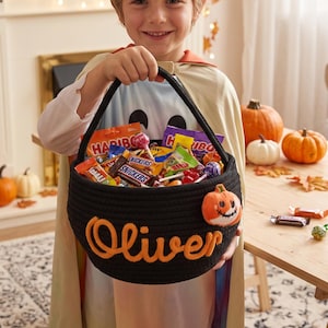 May include: A black woven rope basket featuring the name Oliver embroidered in orange script lettering on the front, accented with a small plush pumpkin patch, filled with an assortment of various Halloween candy and chocolate bars.