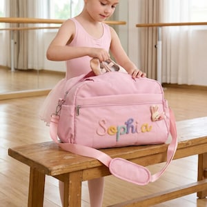 May include: A pink ballet bag with the name "Sophia" embroidered in colourful letters. The bag has a shoulder strap and a small bunny charm. The bag is on a wooden bench.