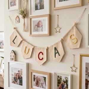 Könnte beinhalten: Eine Wandgalerie mit einem Stoffbanner, das den Namen LUCAS buchstabiert, bestehend aus einzelnen Wimpeln mit jeweils einem farbigen Buchstaben. Das Arrangement ist von mehreren gerahmten Familienfotos, kleinen Holzhaus-Ornamenten und sternförmigen Metalldekorationen umgeben.
