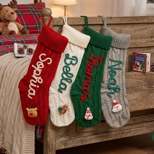May include: Four cable-knit Christmas stockings in red, white, green, and grey. Each stocking features a name embroidered in a contrasting colour: Sophia, Bella, Thomas, and Noah. Small plush holiday icons are attached to the bottom of each.