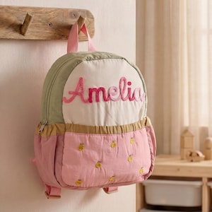 Könnte beinhalten: Ein kleiner, personalisierter Rucksack in Rosa, Grün und Creme, der an einem Holzhaken hängt. Der Rucksack trägt den Namen "Amelia" in Rosa gestickt und hat eine gesteppte Front mit gelben Ananas-Motiven.
