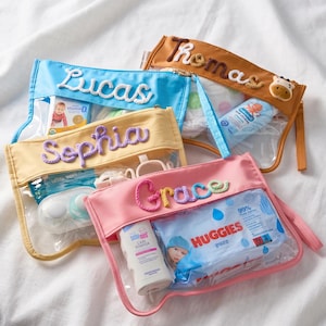 May include: Four clear vinyl pouches with colored fabric borders in blue, yellow, brown, and pink. Each pouch features a personalized name embroidered in yarn: Lucas, Sophia, Thomas, and Grace. The pouches contain various baby care items.