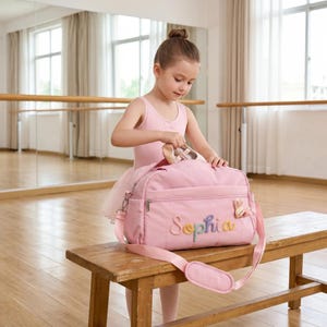 K&ouml;nnte beinhalten: Eine rosa Balletttasche mit dem Namen "Sophia" in bunten F&auml;den gestickt. Die Tasche hat einen Schultergurt und einen kleinen Hasenanh&auml;nger. Die Tasche liegt auf einer Holzbank.