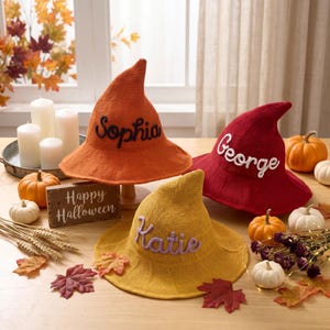 May include: Three knitted witch hats in orange, red, and yellow, each featuring a personalised name embroidered in cursive script: Sophia in black, George in white, and Katie in light purple. The hats have a pointed top and a wide brim.