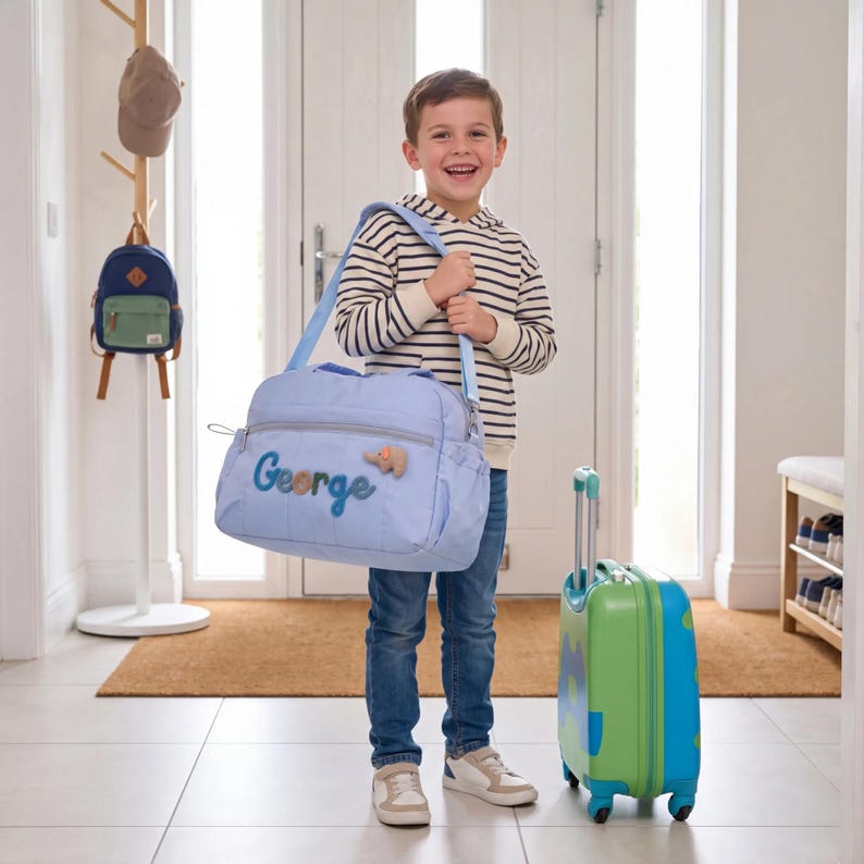 K&ouml;nnte beinhalten: Hellblaue personalisierte Reisetasche mit dem Namen "George" in blauer Schrift gestickt und einem kleinen Elefanten-Applikation. Ein passender blau-gr&uuml;ner Rollkoffer ist ebenfalls vorhanden. Die Tasche hat einen Schultergurt.