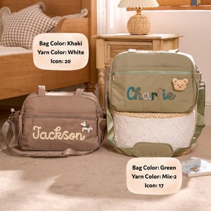 K&ouml;nnte beinhalten: Zwei Wickeltaschen. Eine ist khaki mit dem Namen "Jackson" in wei&szlig;em Garn gestickt und einem Pferde-Symbol. Die andere ist gr&uuml;n mit "Charlie" in Blau gestickt und einem Teddyb&auml;r-Symbol. Beide haben Schultergurte.
