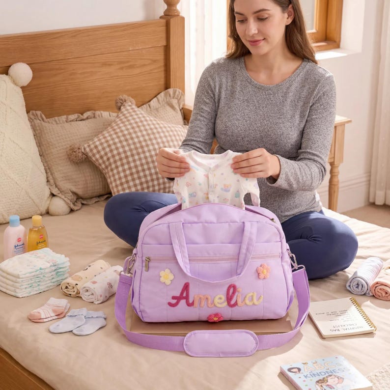 K&ouml;nnte beinhalten: Eine lavendelfarbene Wickeltasche mit dem Namen "Amelia" in bunten F&auml;den gestickt, verziert mit Blumenapplikationen. Ein Baby-Body wird hineingelegt. Weitere Babyartikel, darunter Windeln, Socken und Lotionen, sind in der N&auml;he angeordnet.