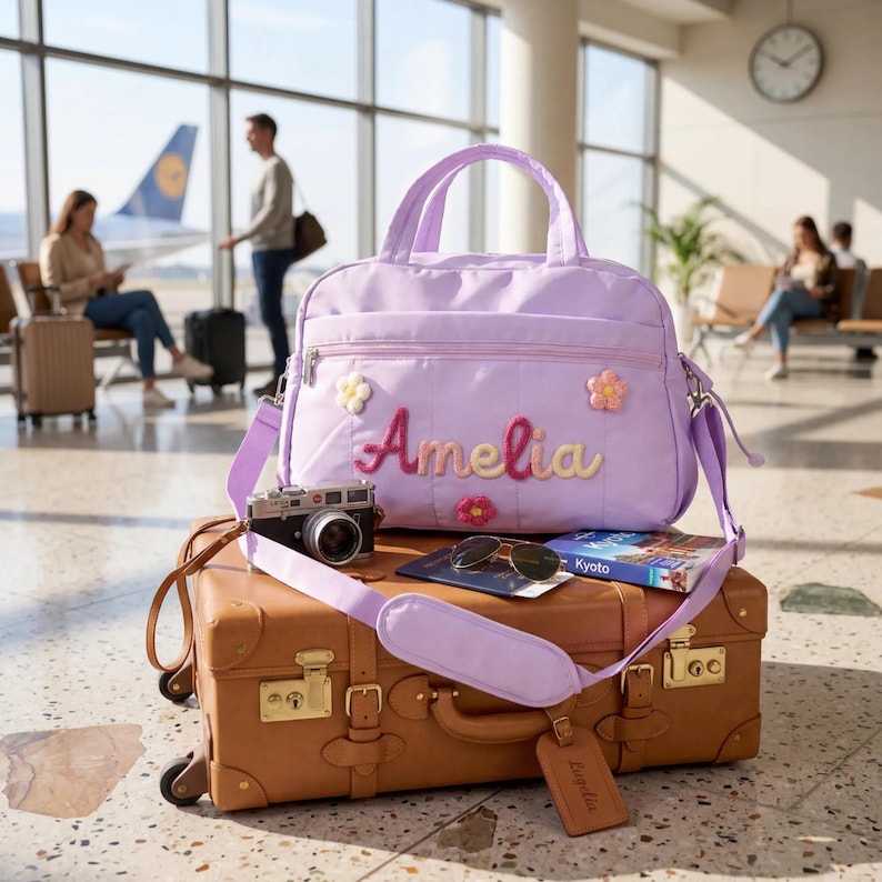 K&ouml;nnte beinhalten: Eine lavendelfarbene Reisetasche mit dem Namen "Amelia" und floralen Verzierungen, platziert auf einem braunen Koffer. Eine Vintage-Kamera, Sonnenbrille und ein Reisef&uuml;hrer sind ebenfalls zu sehen. Die Tasche hat einen Schultergurt und einen passenden Gep&auml;ckanh&auml;nger.