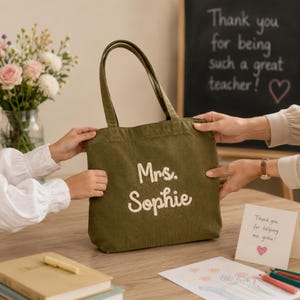 May include: An olive green corduroy tote bag featuring the embroidered white text "Mrs. Sophie" on the front. The bag has two shoulder straps and a rectangular shape. The fabric has a visible vertical ribbed texture throughout the exterior surface.