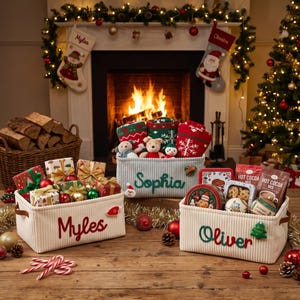 Peut inclure: Trois paniers de rangement rectangulaires en velours côtelé blanc avec des poignées en cuir marron, personnalisés avec les prénoms Myles, Sophia et Oliver brodés en rouge ou vert. Les paniers contiennent des cadeaux, des peluches et du cacao.
