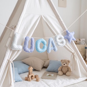 Peut inclure: Une guirlande en tissu épelant le nom LUCAS en lettres rembourrées suspendues à un tipi blanc. Les lettres présentent divers motifs bleus et blancs, notamment du vichy, des rayures et des pois, se terminant par une étoile en vichy bleu.