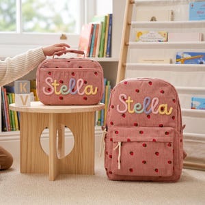 Könnte beinhalten: Ein Rucksack aus altrosa Kord und eine passende rechteckige Lunchtasche, beide mit einem wiederkehrenden Erdbeermuster und dem Namen Stella, der in mehrfarbigen, strukturierten Buchstaben auf der Vorderseite jedes Teils aufgestickt ist.