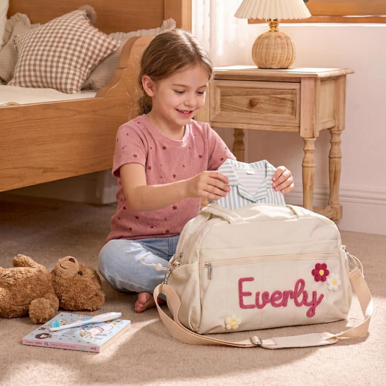 K&ouml;nnte beinhalten: Eine beige Wickeltasche mit dem Namen "Everly" in rosa Schrift und floralen Akzenten. Die Tasche hat einen Schultergurt und ist offen, mit einem gestreiften Hemd darin. Eine junge Person h&auml;lt das Hemd. Ein Teddyb&auml;r und ein Buch liegen auf dem Boden.
