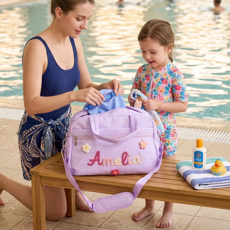 K&ouml;nnte beinhalten: Eine lavendelfarbene Sporttasche mit dem Namen "Amelia" in Rosa und Gelb gestickt. Die Tasche hat einen Schulterriemen und ist mit Blumenapplikationen verziert. Eine Frau und ein Kind bereiten sich auf das Schwimmen vor.