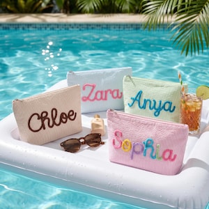 May include: Four rectangular seersucker pouches in beige, light blue, light green, and pink, each featuring a personalised name embroidered in cursive script: Chloe, Zara, Anya, and Sophia. A pair of brown sunglasses and a small bottle sit nearby.