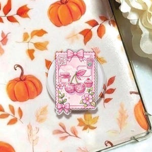 May include: A pink phone grip featuring a cherry and cupcake design. The grip has a scalloped edge and a pink bow at the top. The background has a fall theme with pumpkins and leaves.