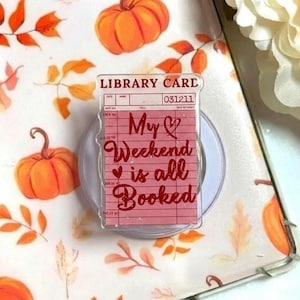 Puede incluir: Un soporte de teléfono transparente con un diseño de tarjeta de biblioteca rosa que dice "My weekend is all Booked". El fondo presenta calabazas naranjas y hojas de otoño sobre una superficie blanca.