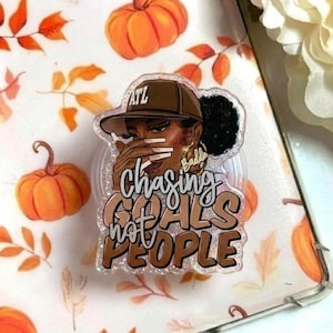 Puede incluir: Un soporte de teléfono acrílico transparente con una mujer que lleva una gorra de béisbol marrón con el texto "ATL". El soporte tiene las palabras "Chasing Goals Hot People" en una fuente brillante. El fondo incluye calabazas naranjas y hojas de otoño.