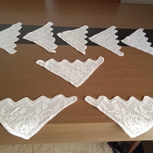 May include: A selection of white, triangular doilies with a scalloped edge, displayed on a wooden surface. The doilies have a delicate, open-weave pattern, suggesting a vintage or handmade origin.