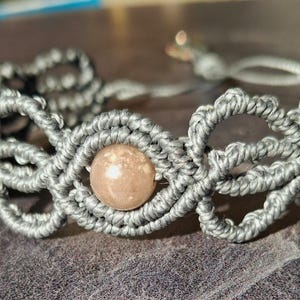 Può includere: Primo piano di un braccialetto macramè grigio con una perla centrale rotonda marrone chiaro. L'intricato design intrecciato del braccialetto è visibile, con una delicata chiusura. Il braccialetto è esposto su una superficie strutturata.
