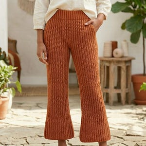 May include: Rust-coloured knitted trousers with a flared leg. The trousers are made of a ribbed knit fabric and are worn with a white blouse. The background features potted plants and a stone patio.