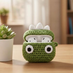 May include: A green crocheted dinosaur-themed case for wireless earbuds, with white accents and large black and white eyes. The case is open, revealing white earbuds inside. A small succulent in a white pot is to the left.