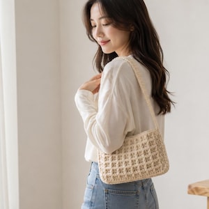 May include: A beige, woven shoulder bag with a rectangular shape and a matching strap. The bag has a grid-like pattern and is worn by a person wearing a white long-sleeve shirt and blue jeans. The background is a neutral white wall and a wooden table.
