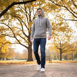 May include: A gray hooded sweater with a textured knit pattern and a front pocket. The person is wearing dark blue jeans and white sneakers. The background features a park with trees and fall foliage.