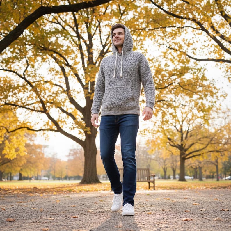 May include: A gray hooded sweater with a textured knit pattern and a front pocket. The person is wearing dark blue jeans and white sneakers. The background features a park with trees and fall foliage.