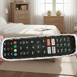 May include: A crochet remote control holder in lavender and white. The holder is designed to fit a standard remote, with a rectangular shape and a decorative edge. The holder is held in front of a bedroom setting.