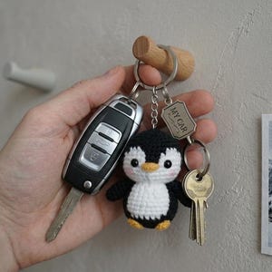Może przedstawiać: Brelok do kluczy z czarnym i białym kluczykiem samochodowym, małym szydełkowym pingwinem i kluczem z zawieszką "MY CAR". Pingwin ma żółty dziób i stopy. Brelok wisi na drewnianym haczyku.