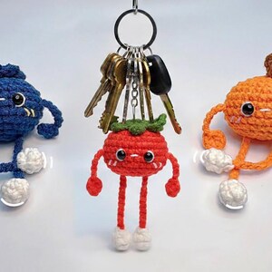 May include: Three crocheted fruit-shaped keychains: a blue blueberry, a red tomato, and an orange. Each has arms, legs, and white accents. The keychains are attached to a keyring with several keys.