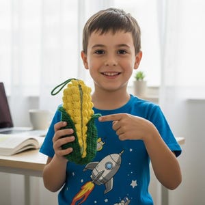 May include: A yellow and green crocheted corn-shaped pouch with a zipper closure and a green loop. The pouch is held by a person wearing a blue t-shirt with a rocket ship graphic.