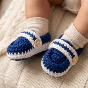 May include: A pair of handmade baby shoes in a nautical style. The shoes are crocheted with a deep blue yarn and accented with white stripes and trim. Each shoe features a small wooden button closure.