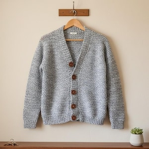Op de afbeelding: Een grijze gebreide cardigan met een V-hals en houten knopen hangt aan een houten hanger. De trui wordt gepresenteerd tegen een neutrale muur. Een kleine vetplant in een witte pot staat op een houten oppervlak.