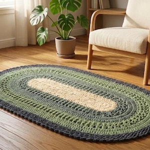 Può includere: Un tappeto ovale fatto a mano con un design all'uncinetto. Il tappeto presenta una sezione centrale beige, circondata da strati di verde e grigio scuro. Il tappeto è posizionato su un pavimento in legno, vicino a una finestra, una sedia e una pianta in vaso.