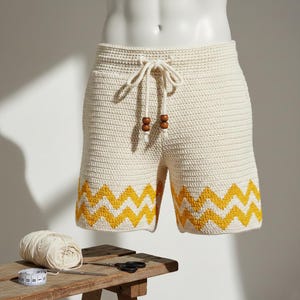 May include: Cream crochet shorts with a yellow zigzag hem. The shorts feature a drawstring waist with wooden bead details. The garment is displayed on a mannequin, highlighting the handmade texture and design. The shorts are approximately 40 cm in length.