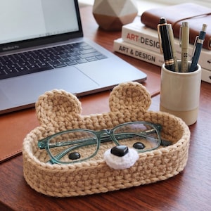 May include: A handmade, light brown crochet eyeglasses holder shaped like a bear. The holder features two small ears and a white and black nose. A pair of green-framed glasses rests inside the holder, which sits on a wooden desk.