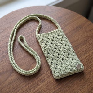 May include: A light sage green crochet phone pouch featuring a textured shell stitch pattern and a long matching strap. The rectangular pouch includes a single silver-toned metal snap button closure at the bottom right corner for securing items.