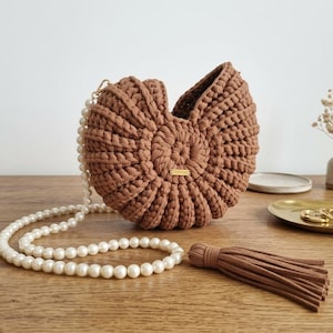 Può includere: Una borsa marrone all'uncinetto con una tracolla a collana di perle. La borsa ha un design a conchiglia e una piccola etichetta color oro. Sono visibili anche un nappa marrone e gioielli color oro.