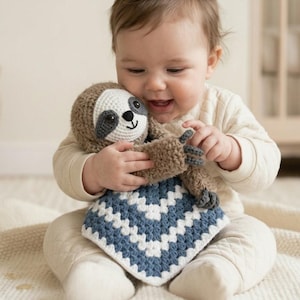 May include: A smiling baby holds a handmade crochet sloth toy with a blue and white chevron patterned blanket. The sloth has a brown body, a white face, and gray accents. The baby is wearing a cream-colored outfit.