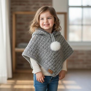 Puede incluir: Un poncho de ganchillo gris con capucha, con un patrón texturizado y un pompón blanco. El poncho tiene una pequeña etiqueta marrón y lo lleva un niño. El niño lleva una camiseta blanca de manga larga y vaqueros azules.