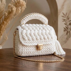 Puede incluir: Un bolso blanco tejido a mano con un diseño trenzado en la solapa delantera. El bolso presenta una correa y asa de cadena dorada, un cierre dorado y una borla. El bolso está sobre una superficie de madera.