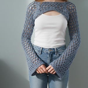 May include: A blue crochet shrug with long sleeves and a keyhole opening. The open weave design of the shrug is visible. The model is wearing a white top and blue jeans. The shrug is a unique fashion accessory.