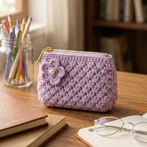 Pode incluir: Uma bolsa de crochê lavanda com um padrão texturizado de pontos concha e um fecho de zíper dourado. Uma pequena flor de crochê de seis pétalas com uma pérola branca central está fixada no lado frontal esquerdo do acessório retangular.